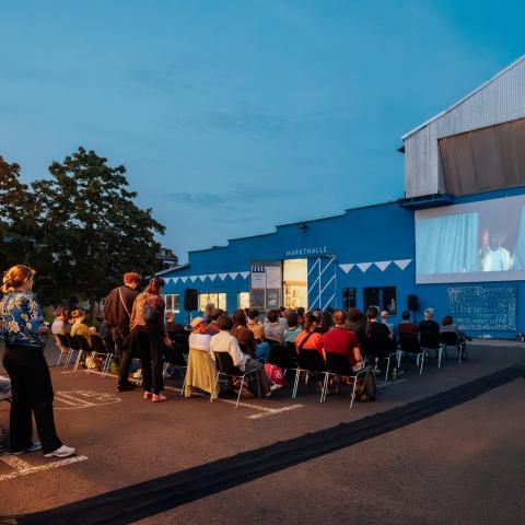 Open-Air-Kino am Abend: Etwa 50 Personen schauen gemeinsam in stimmungsvoller Beleuchtung einen Film, der an die Wand einer Markthalle projiziert wird.