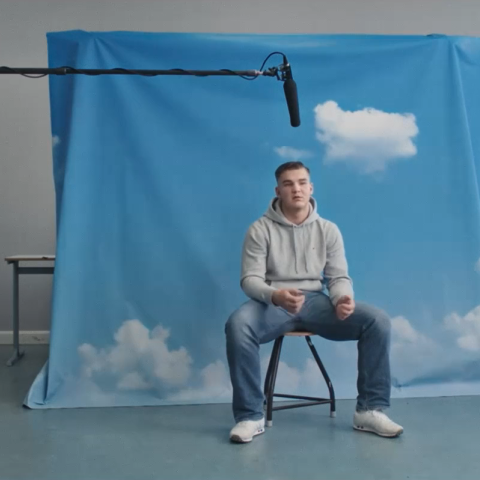 Jugendlicher sitzt vor aufgestelltem Wolken-Hintergrund. Über ihm hängt ein Mikrofon.