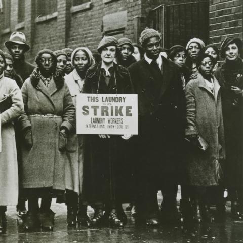 A group of laundry workers stand on the street during a strike in the 1930s, holding signs.