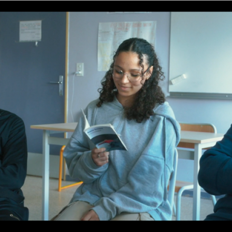 A group of young people sits in a classroom in a circle; one person in the middle is reading from a booklet while the others listen attentively.