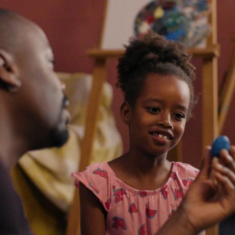 Ein Schwarzes Mädchen sitzt neben einem Schwarzen Mann und schaut grinsend auf ein kleines blaues Ei, das er hochhält