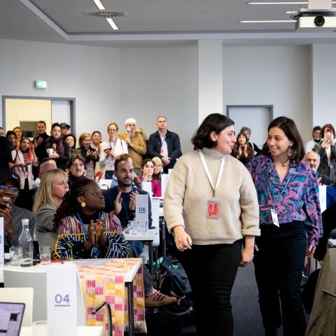 A business room full of applauding and smiling people. Two young, smiling women are making their way through the crowd.