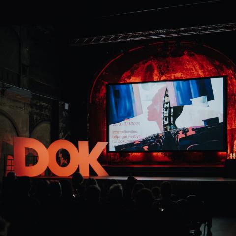 Publikum sitzt vor einer Bühne mit einer Leinwand, die den Festivaltrailer von DOK Leipzig zeigt; links auf der Bühne ist in roten Großbuchstaben der Schriftzug DOK zu sehen