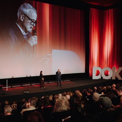 Two people stand on stage before a red curtain and large audience at DOK Leipzig; one speaks into a microphone, and the scene is projected on the big screen.