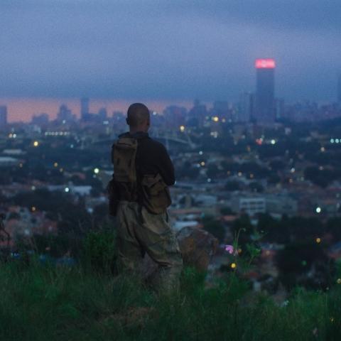 A bald person stands on a hill at dusk, looking out over a city with glowing high-rise buildings. They are wearing camouflage pants and several small pouches on their body.