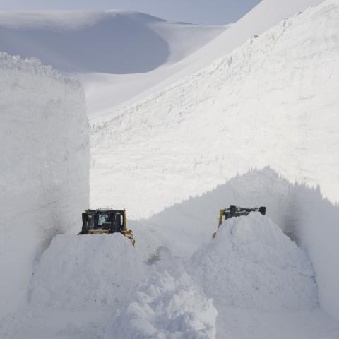 Two snowplows clear a deep, narrow path through towering snow in a snowy mountain landscape.