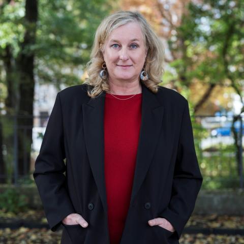 Eine Person mit blonden, schulterlangen Haaren steht im Freien vor Bäumen, trägt einen schwarzen Blazer über einem roten Oberteil und blickt leicht lächelnd direkt in die Kamera.