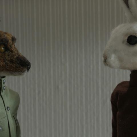Zwei Stofffiguren, ein Fuchs in grüner Jacke und ein Hase in braunem Pullover, stehen sich vor einer neutralen Wand gegenüber.