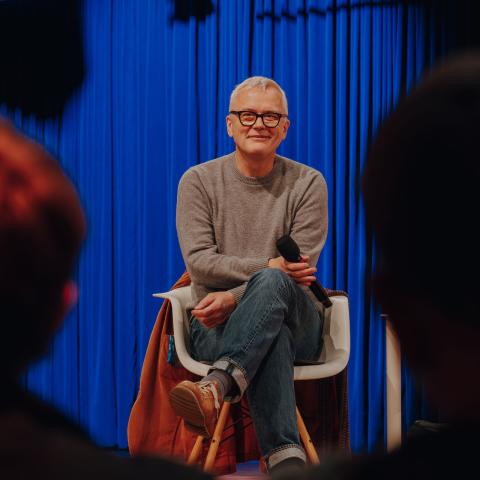 Man with gray hair and glasses sits smiling with a microphone in front of a blue curtain, audience in the foreground.