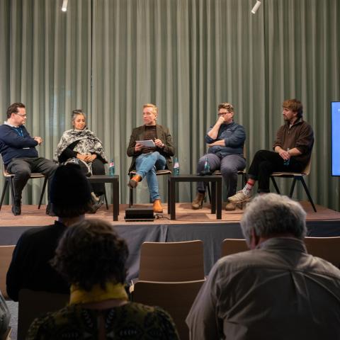 Eine moderierte Podiumsdiskussion mit fünf Personen auf einer Bühne vor Publikum. Neben der Bühne steht ein Bildschirm mit der Aufschrift „Welcome to DOK Exchange XR“.