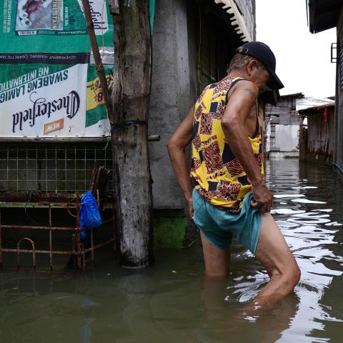 A man is wading through knee-deep water that has flooded a street between closely packed houses.