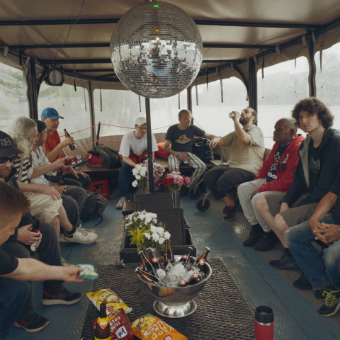 Eine Gruppe Männer sitzt auf einem Boot, von der Decke hängt eine Diskokugel und darunter steht ein Beistelltisch mit Snacks und gekühltem Bier.