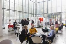 Blick in das Festivalzentrum von DOK Leipzig: Ein hoher Raum mit gläsernen Wänden, darin mehrere Personen im Gespräch an Kaffeetischen, im Hintergrund der Gästecounter mit mehreren Personen, die dort anstehen.