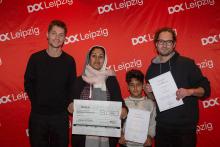 Vier Personen beim Fotocall vor einer DOK Leipzig Logowand: Die beiden Regisseure des Films "Nasim" rahmen die Protagonistin Nasim mit ihrem Sohn ein. Sie halten zwei Preisurkunden in die Kamera und lächeln alle.