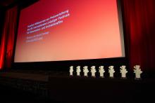 Acht Preis-Statuetten der DOK Leipzig Tauben stehen aufgereiht unterhalb einer Kinoleinwand. Auf der Leinwand steht: Herzlich Willkommen zur Preisverleihung des 64. Internationalen Leipziger Festivals für Dokumentar- und Animationsfilm. 