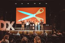 Sieben Personen stehen auf einer Bühne und halten Preisstatuetten in die Höhe. Im Hintergrund das Festivalmotiv von DOK Leipzig 2023: eine riesige Schere.