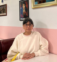 An Asian brown woman sits on a burgundy sofa, wearing a white pullover, medium brown-rimmed glasses, and gold hoop earrings. Her fringe grazes the top of her glasses, and her hands rest on the table. Behind her, a pink lower wall is adorned with mirrors and film posters.