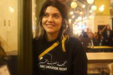 A medium shot of a woman in her thirties, smiling and looking off camera. She has brown eyes and dark brown hair with grey streaks which is tied back. She is wearing a black sweatshirt that reads ’sonic liberation front’ and sits against a window frame