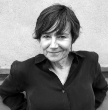 Black-and-white portrait of a smiling woman with short, tousled hair, wearing a dark button-up shirt and standing against a plain textured wall.