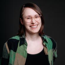 A smiling woman with shoulder-length brown hair and glasses, wearing a green and black patterned shirt against a dark background.