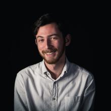 A medium shot of a man smiling and looking into the camera. He has dark hair, a red-ish beard and a white button-down shirt.