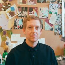 Portrait of a person in a black shirt, photographed indoors in front of a colourful bulletin board covered with drawings, photographs and notes.