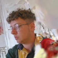 Side-profile portrait of a person wearing round glasses and a colourful jacket, photographed indoors against a bright, softly blurred background.