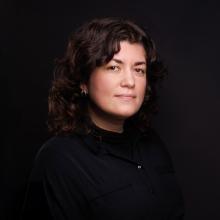 A portrait of a white woman in her thirties. She has brown eyes and curly brown hair. She is wearing a black shirt and is photographed against a black background.