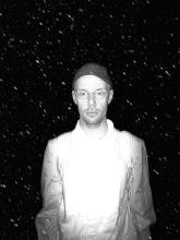 Black-and-white portrait of a person wearing round glasses, a cap and light-coloured clothing, standing against a dark background filled with bright specks.