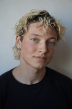 Eine Nahaufnahme eines Mannes in den Zwanzigern, der leicht lächelt und in die Kamera blickt. Hinter ihm ist eine weiße Wand zu sehen. Er hat blaue Augen und kurzes blondes Haar mit dunklen Haarwurzeln. Er trägt ein schwarzes T-Shirt. 