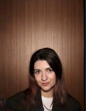 Eine Nahaufnahme einer brünetten Frau mit braunen Augen, die in die Kamera blickt. Ihr Haar ist offen und glatt, und sie trägt eine schwarze Jacke mit einem weißen Oberteil darunter. 