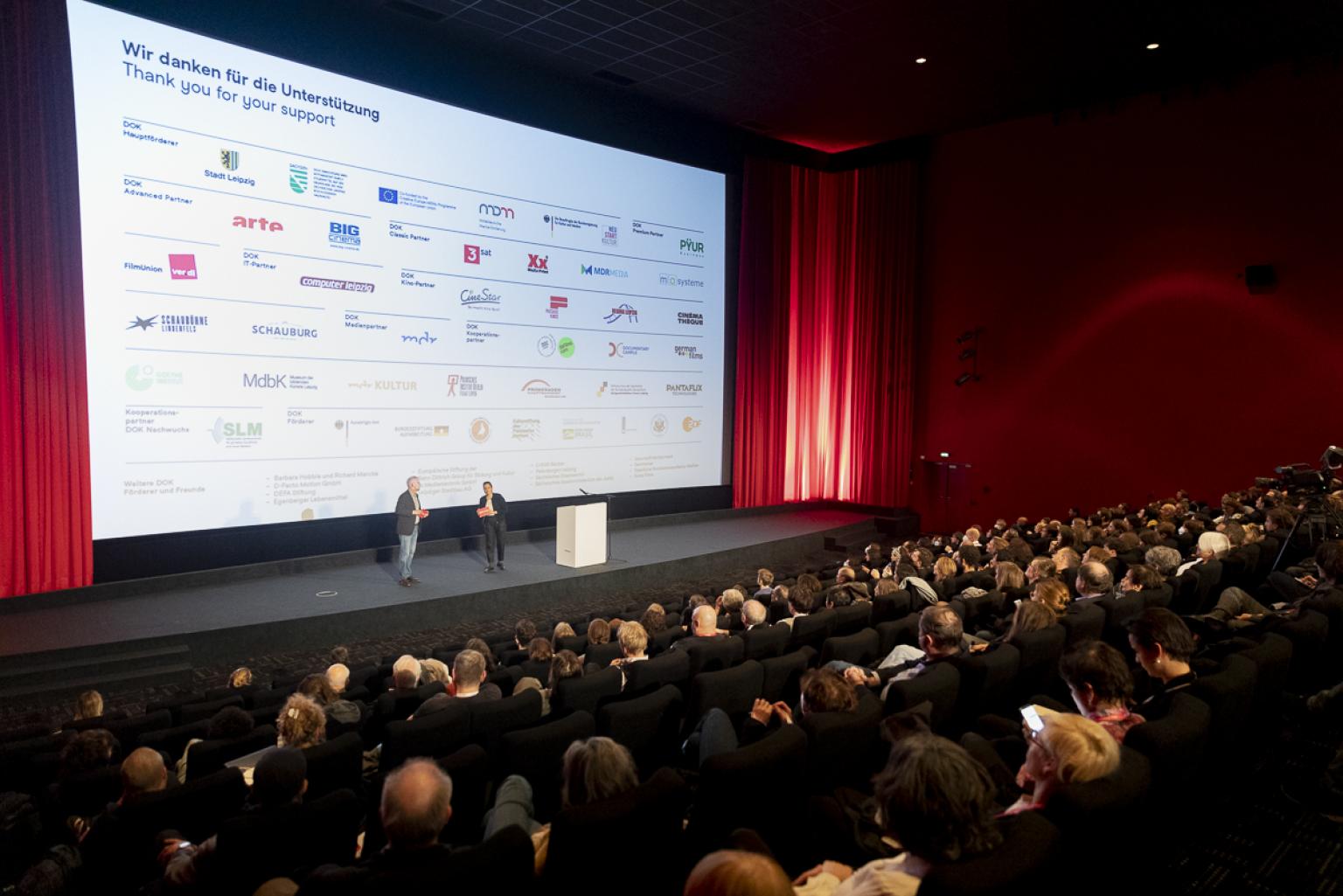 Blick über einen vollbesetzten Kinosaal auf eine Bühne vor der Leinwand. Die Leinwand zeigt eine Übersicht der Sponsoren und Partner von DOK Leipzig.