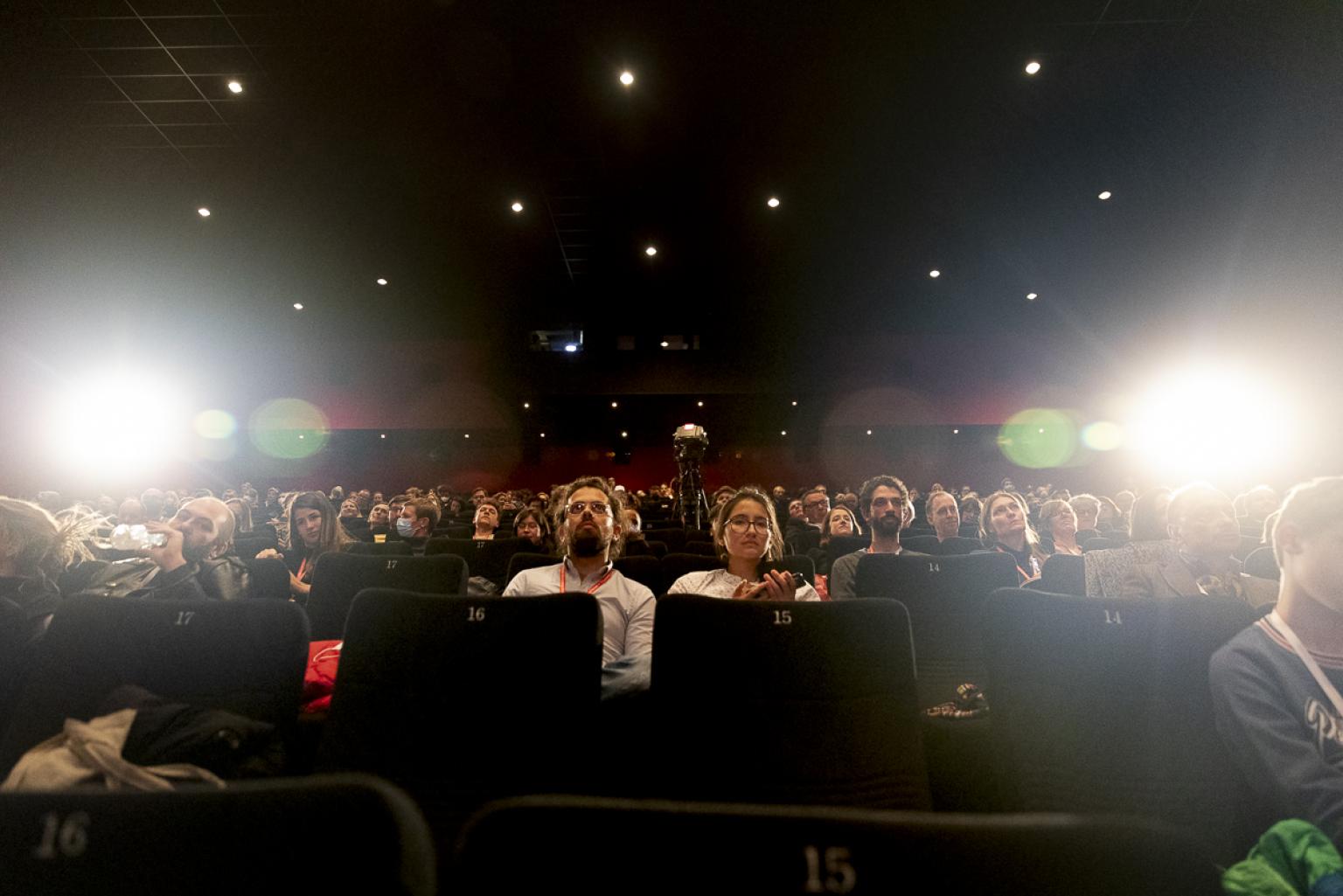 Blick auf die aufsteigenden Sitzreihen eines Kinosaals im Gegenlicht. Fast alle Plätze sind belegt.