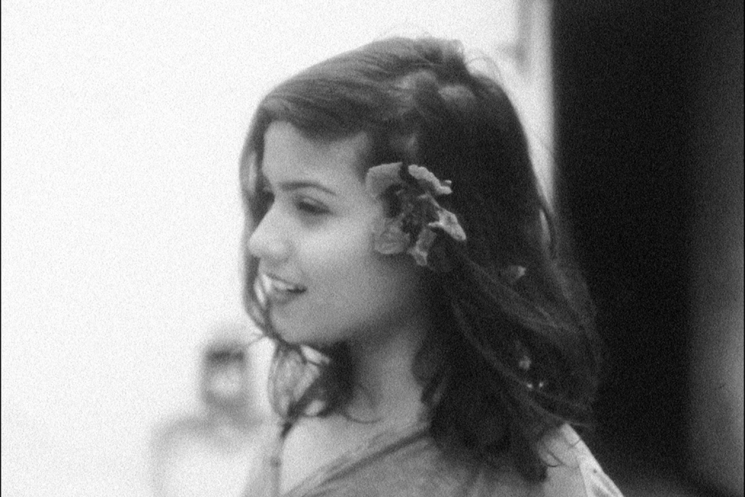 Eine schwarz-weiß Aufnahme: Eine junge Frau im Profil. Sie lächelt und hat sich eine große Blume hinter ihr Ohr gesteckt. Die schulterlangen dunklen Haare trägt sie offen.
