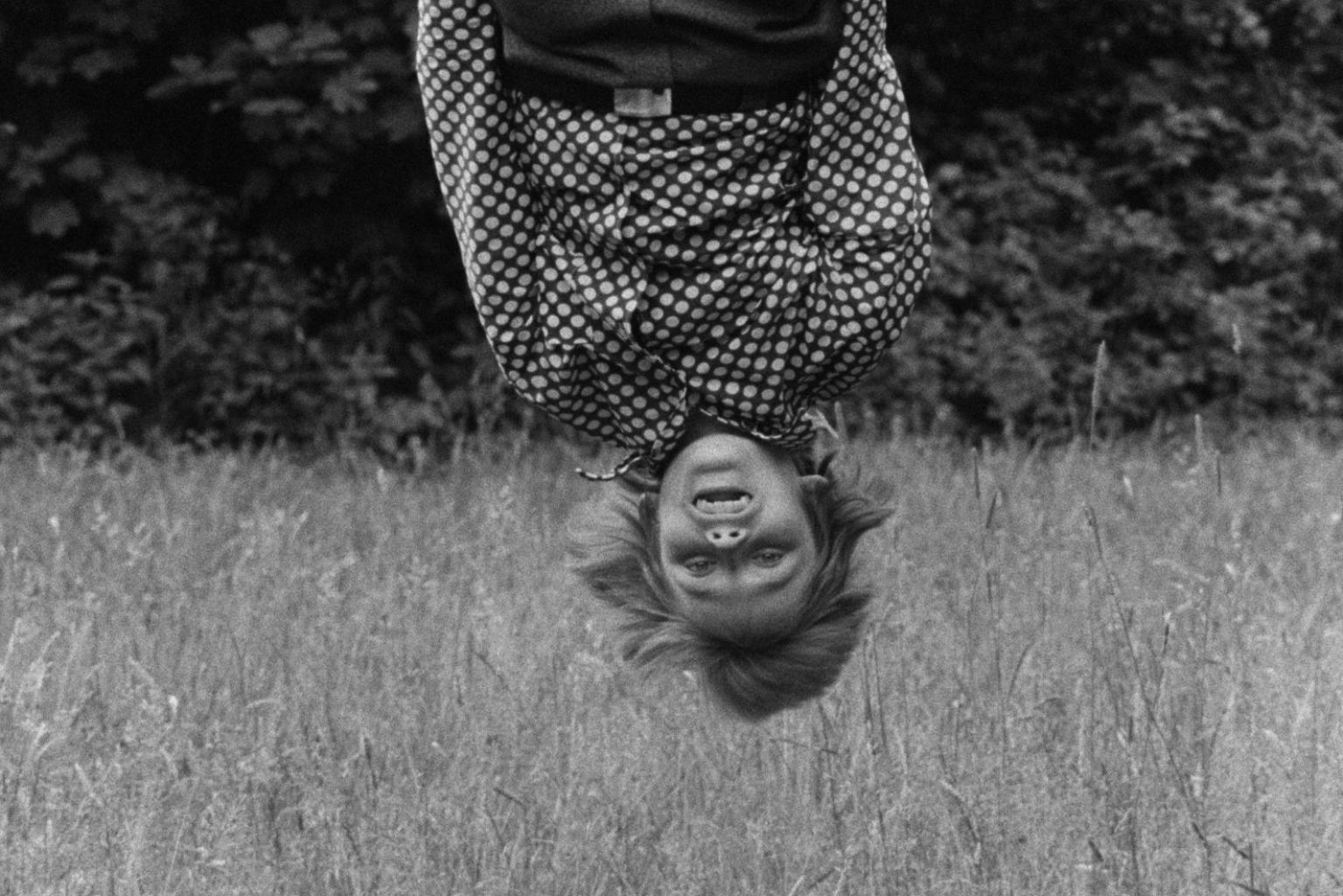 A black and white image of a young person hanging head over heels into the frame, with a cornfield in the background. The person is wearing a polka-dotted shirt and is smiling into the camera.
