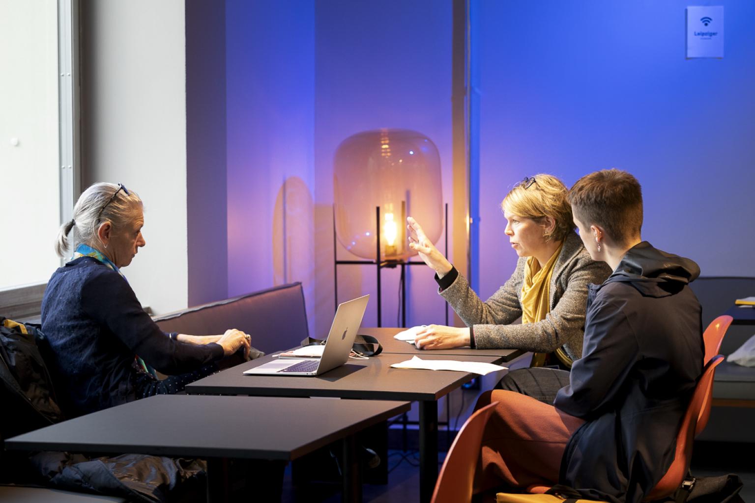 Drei Frauen sitzen in produktiver Atmosphäre zusammen an einem Tisch und unterhalten sich. Ein Laptop steht auf dem Tisch. Die Person in der Mitte gestikuliert, die beiden Personen links und rechts neben ihr schauen sie interessiert an.