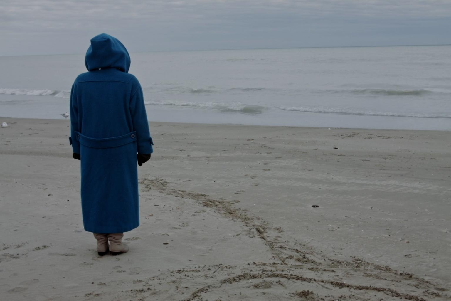 Am Strand, vor dem flachen, ruhigen Meer, steht eine Person in beinahe bodenlangem blauen Regenmantel mit dem Rücken zur Kamera. Sie hat die riesige Kapuze hochgezogen und schaut zum Meer.