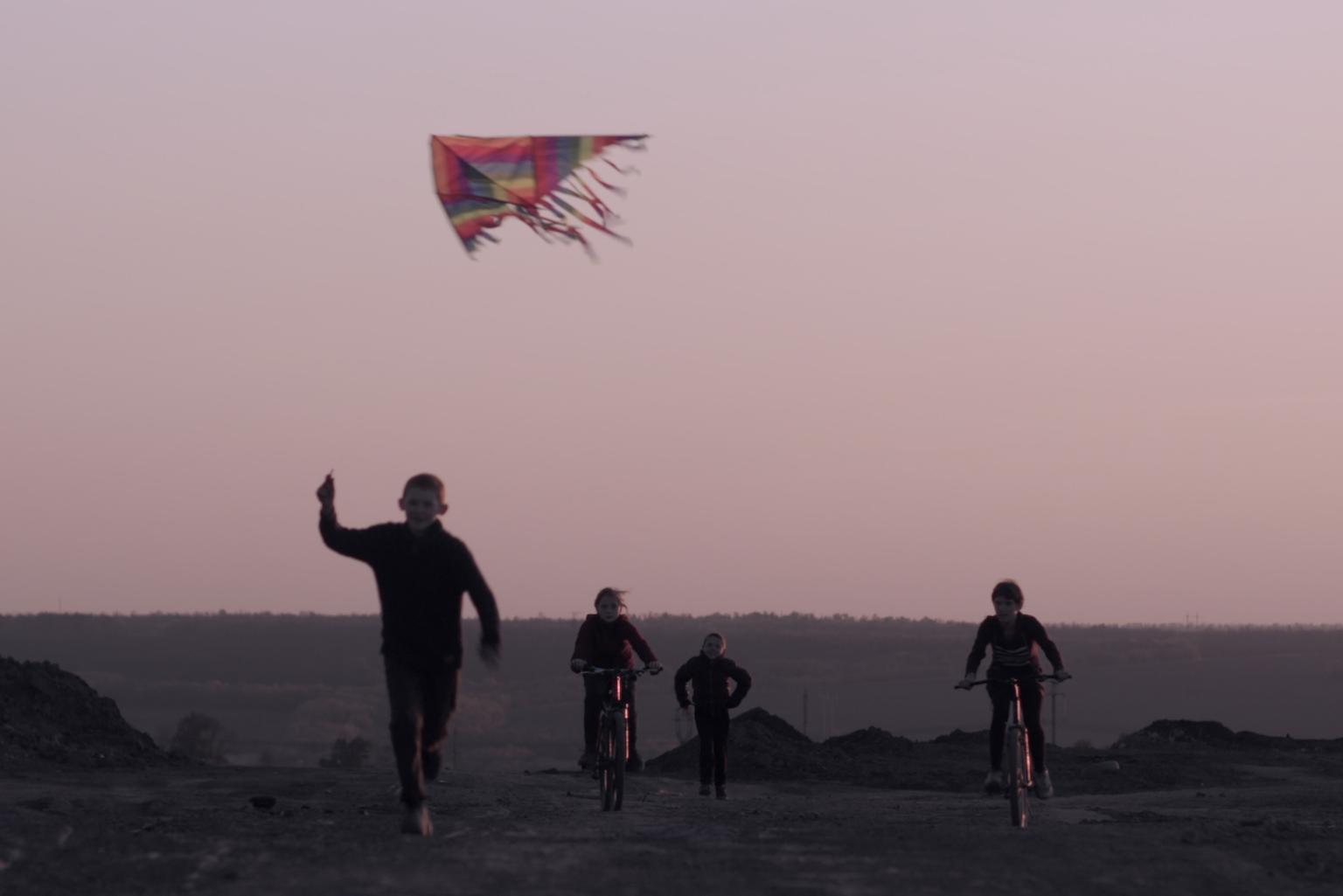 A wide area or street at dusk: four children move towards the camera. Two are running, two are riding their bicycles. Above them flies a kite, which the child on the far left holds by the string.
