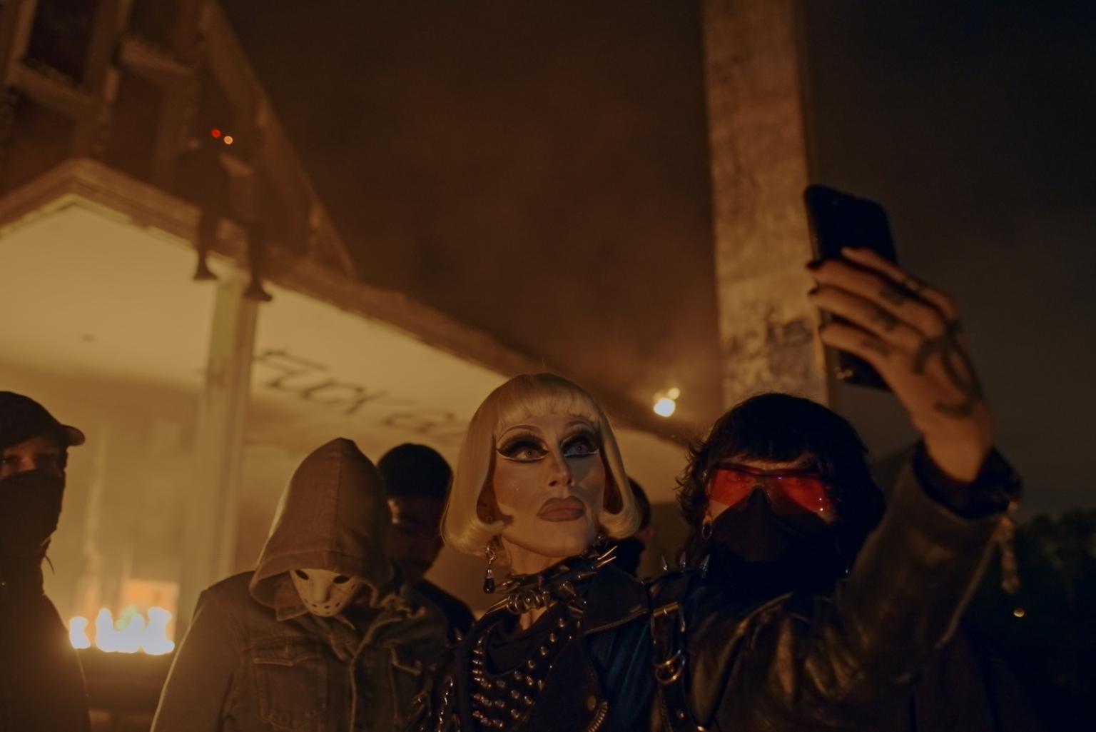 In the center you can see a heavily made-up person with striking facial features. She wears a white-blond wig with a bob cut and black patent clothing. She is taking a selfie with a serious expression. To her left and right are people wearing face masks and hoods. Everything looks a little sinister, but also rebellious and powerful.