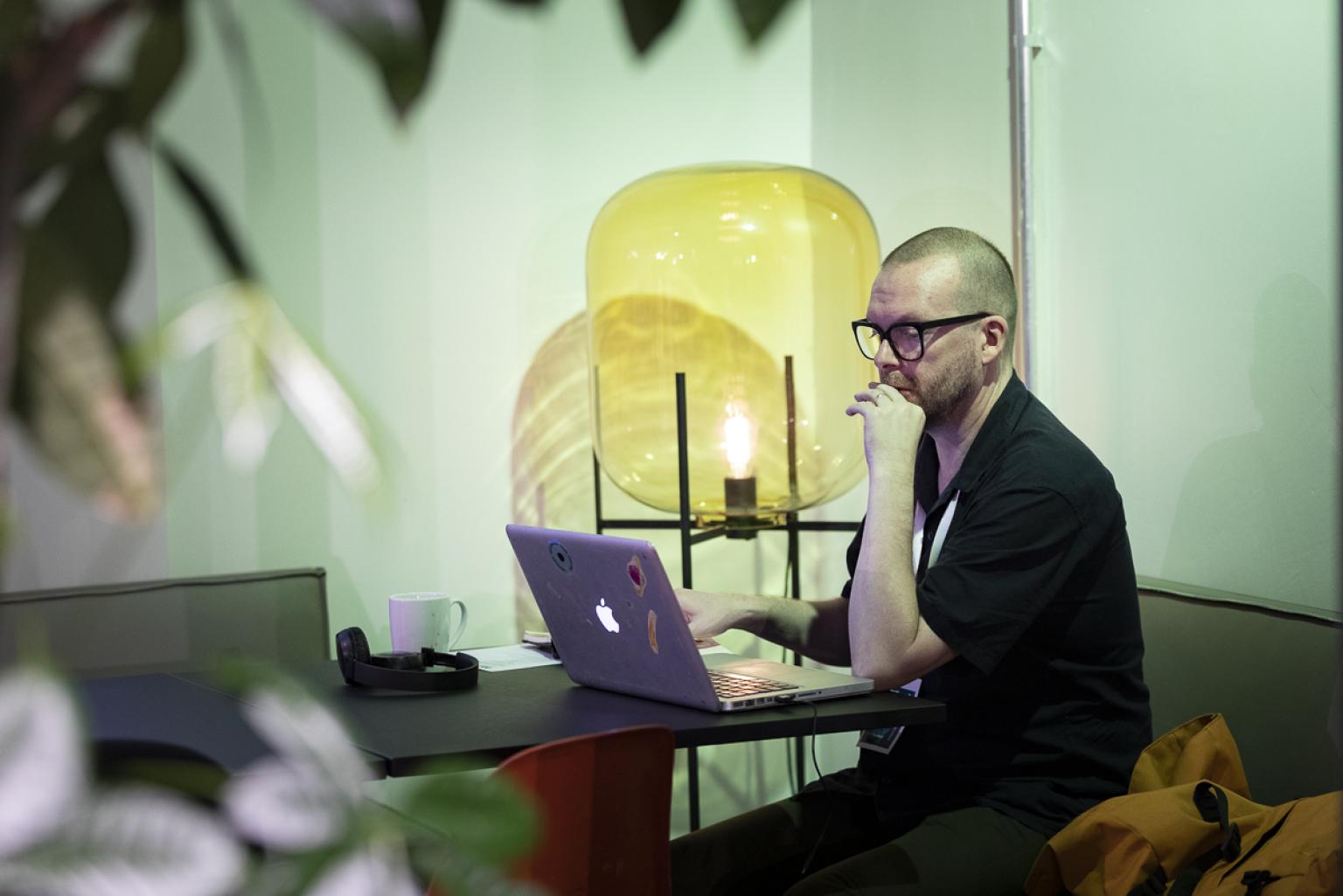 A man sits at a table framed by plants and a giant yellow glass lamp, looking at his laptop.