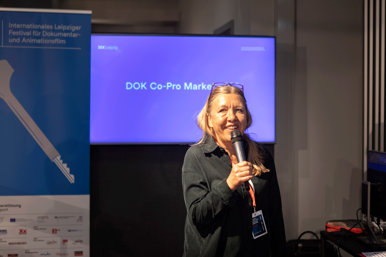 Director of DOK Industry Nadja Tennstedt stands in a room, talking into a microphone. In the background a screen with text: "DOK Co-Pro Market".