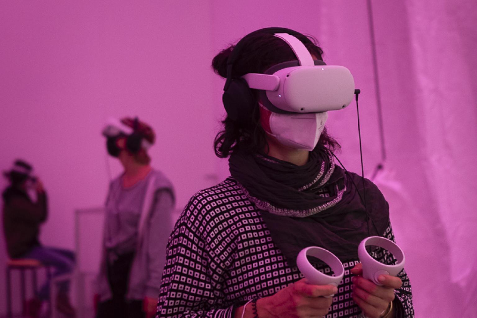 A woman wearing a VR head set and holding two hand controllers in her hands stands in a pink room. In the background another person with a VR head set.