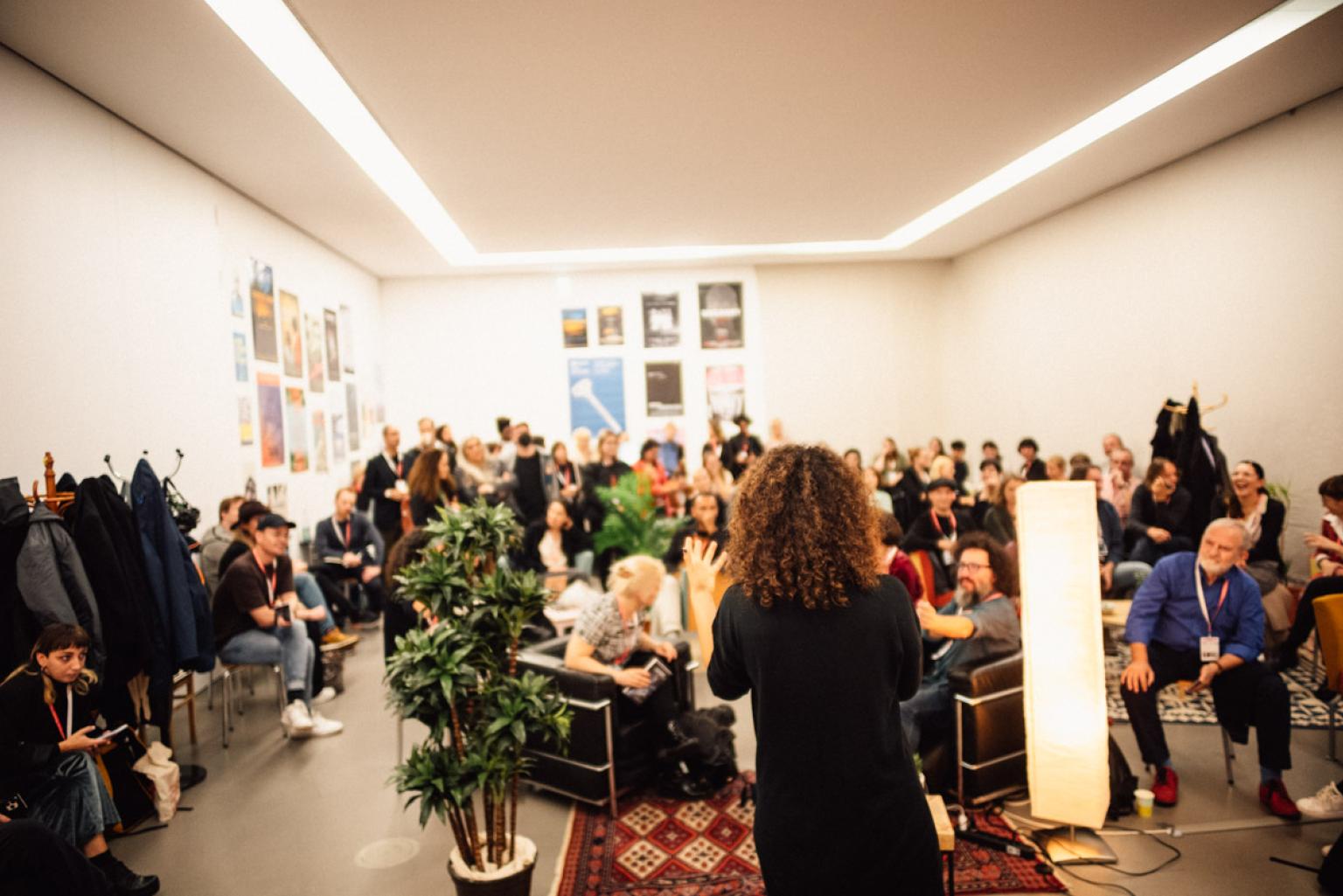 A room full of people who sit and stand while listening to a person who is speaking into a microphone.
