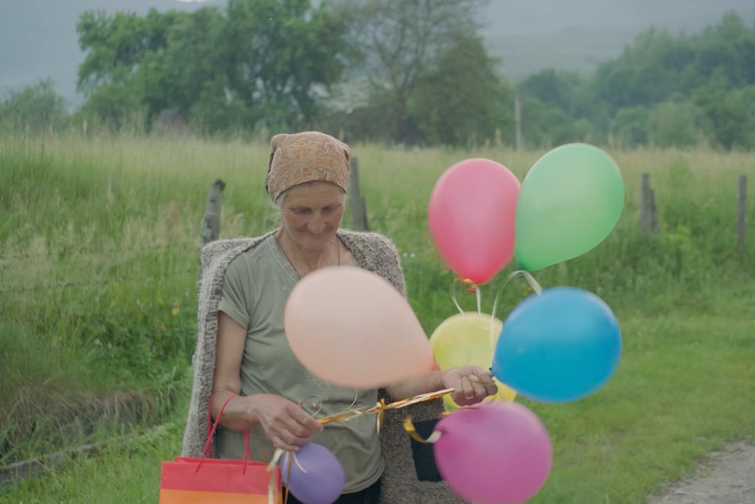 Eine ältere Frau blickt lächelnd auf mehrere bunte Luftballons, die sie in der Hand hält.