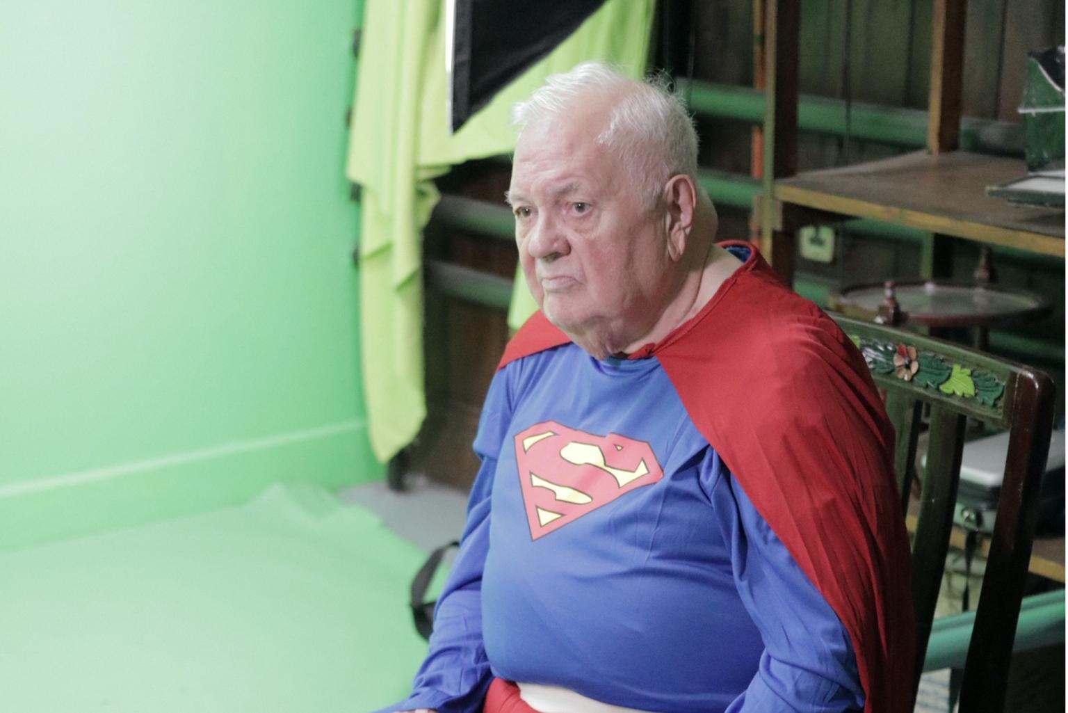 Ein älterer Mann sitzt in einem Superman-Kostüm vor einer grünen Leinwand in einem Fotostudio.