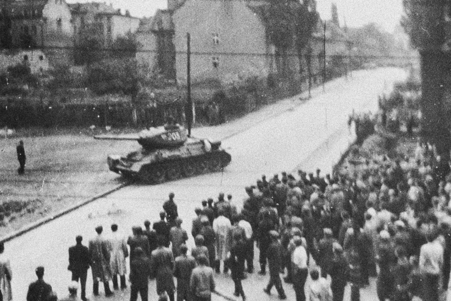 Schwarz-Weiß-Aufnahme: Eine Menschenmenge bewegt sich auf einen Panzer zu, der an einer Straßenecke steht.