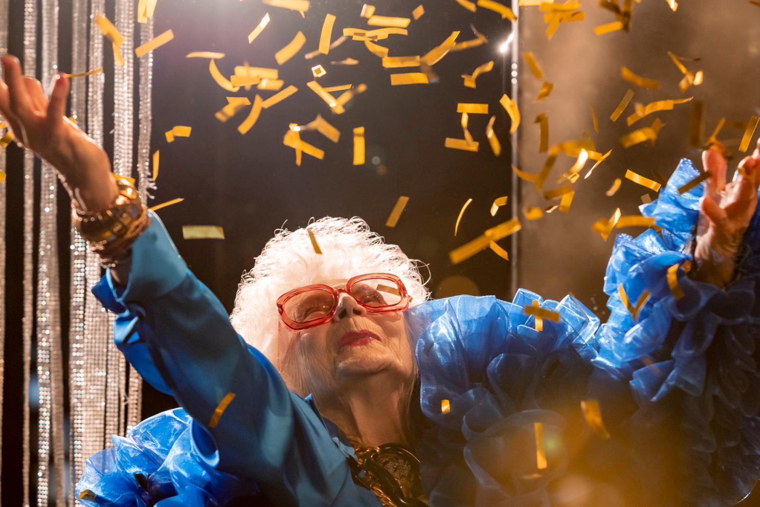 Older lady with oversized red glasses, blue costume and many frills throws cocky confetti in the air in front of glitter curtain