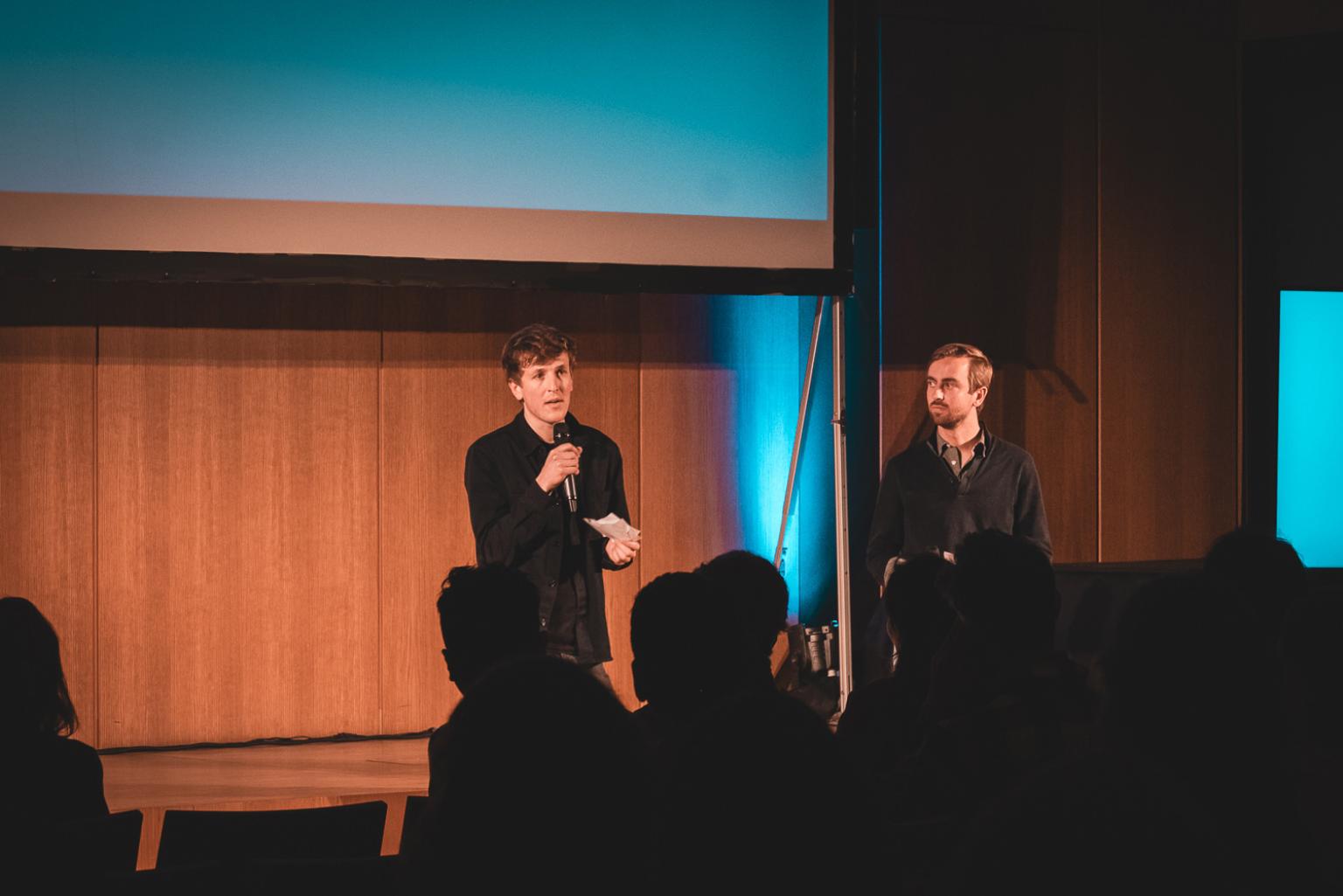 Two persons speak to a seated audience in a wooden-paneled room.