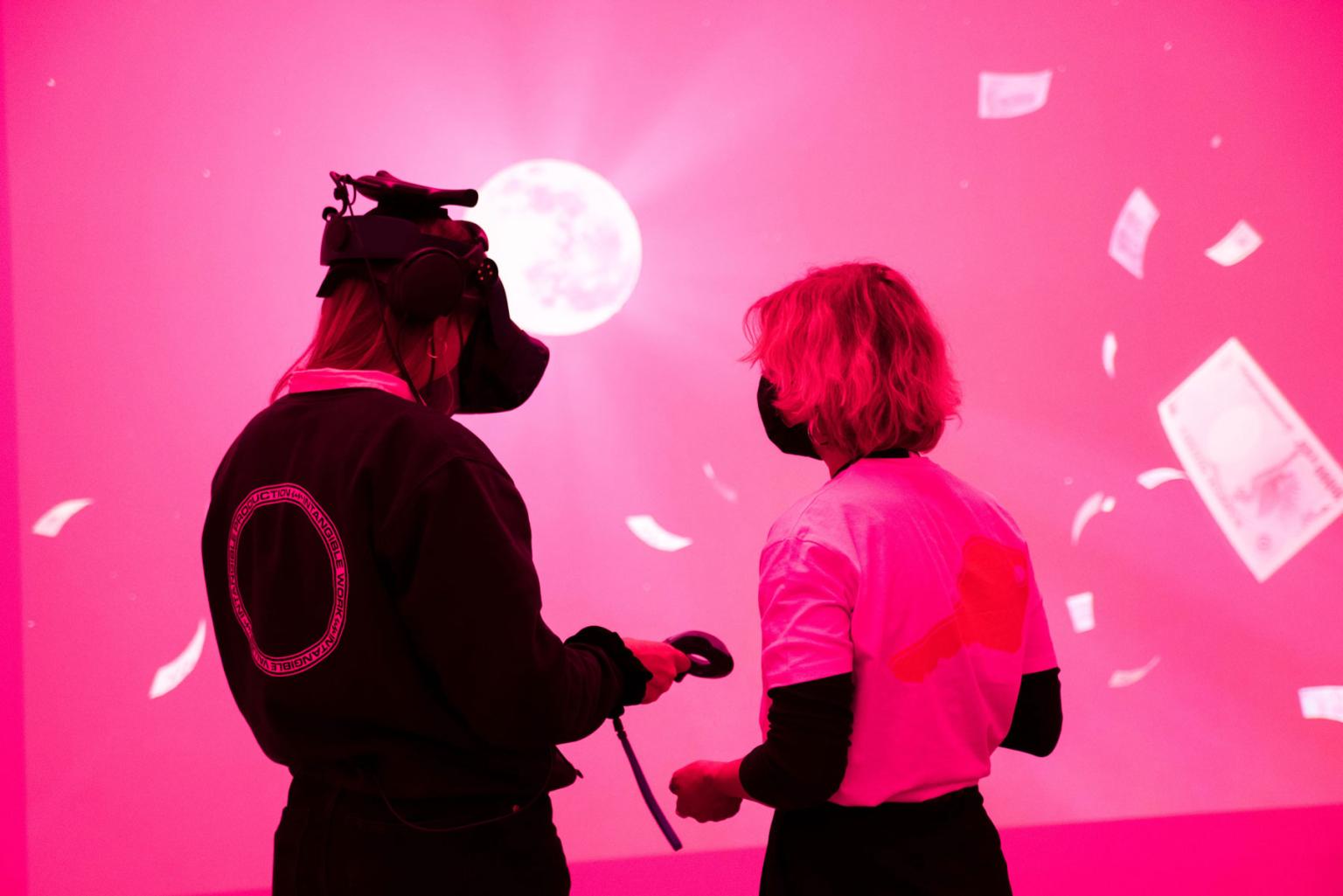 Zwei Personen stehen in einem Raum, der in Pinkes Licht getaucht ist. Vor ihnen eine riesige Leinwand, auf der ein Mond und fliegende Geldscheine zu sehen sind. Die Person links trägt eine VR-Brille.
