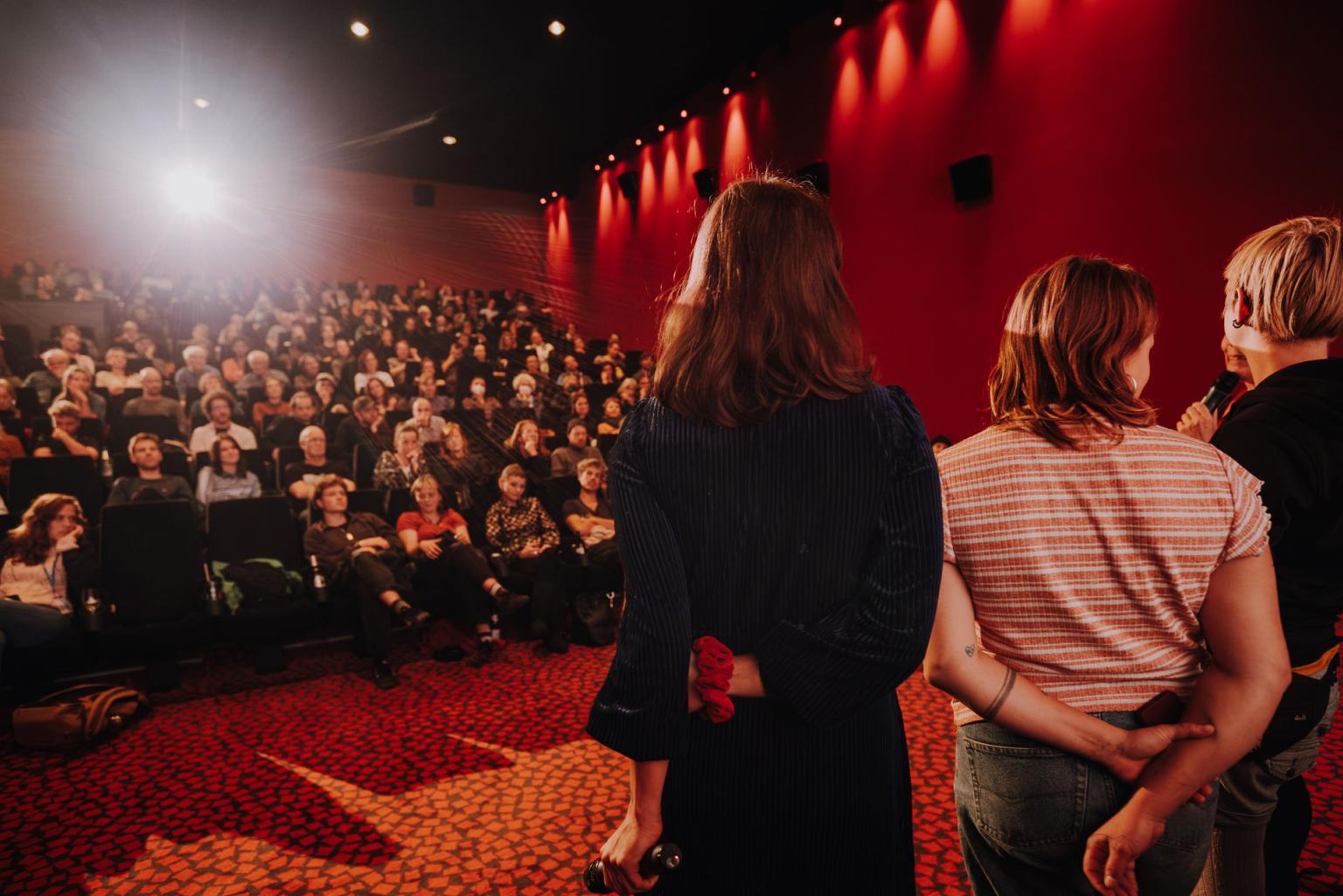 Im Kino stehen drei Menschen auf der Bühne vor einem vollen Saal im strahlenden Licht der Projektion